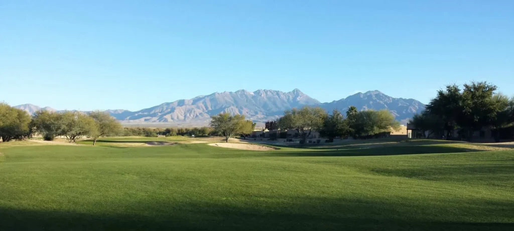A wide, well-manicured fairway stretches toward a bunker-flanked green, framed by desert trees and dramatic mountain ranges under a clear blue sky — the defining landscape of desert golf in Arizona.