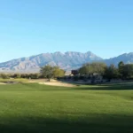 A wide, well-manicured fairway stretches toward a bunker-flanked green, framed by desert trees and dramatic mountain ranges under a clear blue sky — the defining landscape of desert golf in Arizona.