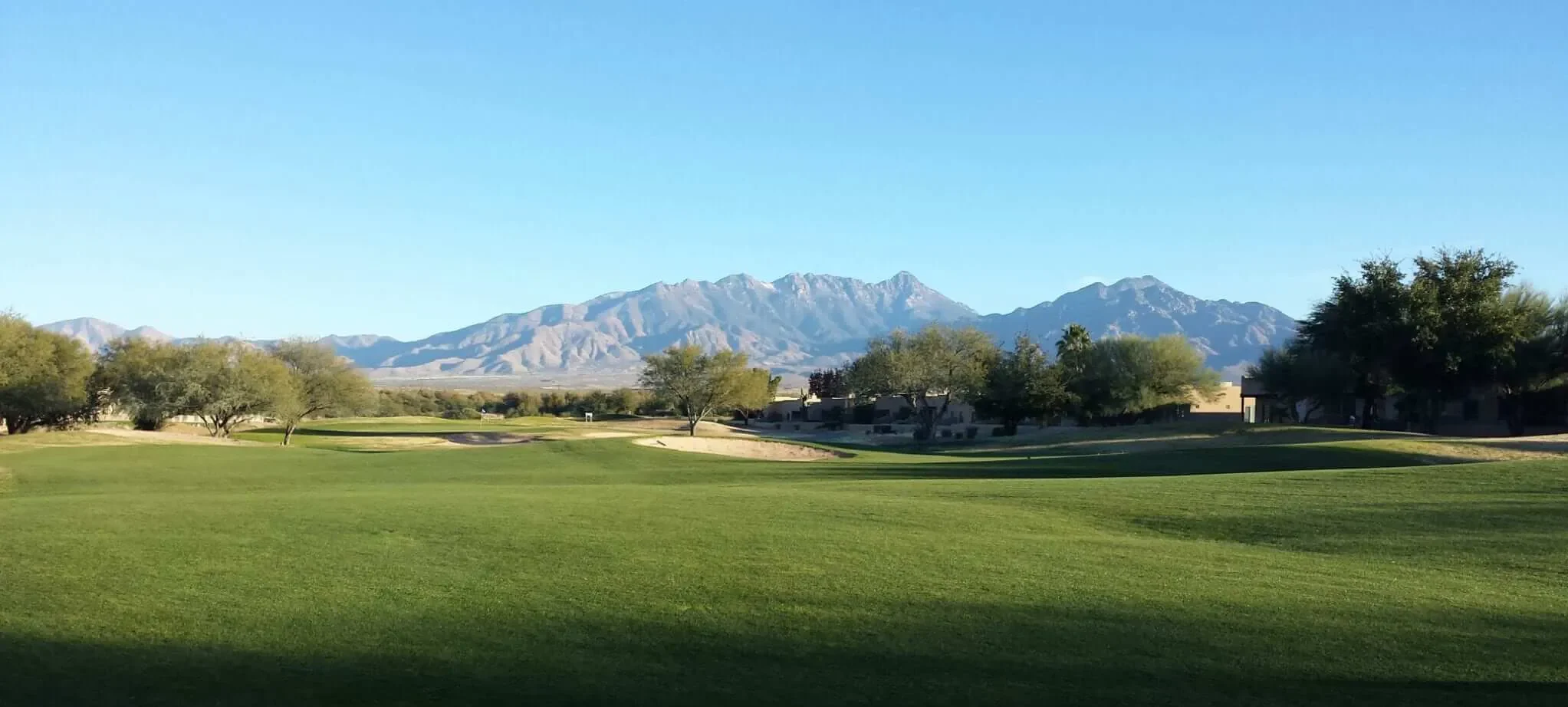 A wide, well-manicured fairway stretches toward a bunker-flanked green, framed by desert trees and dramatic mountain ranges under a clear blue sky — the defining landscape of desert golf in Arizona.